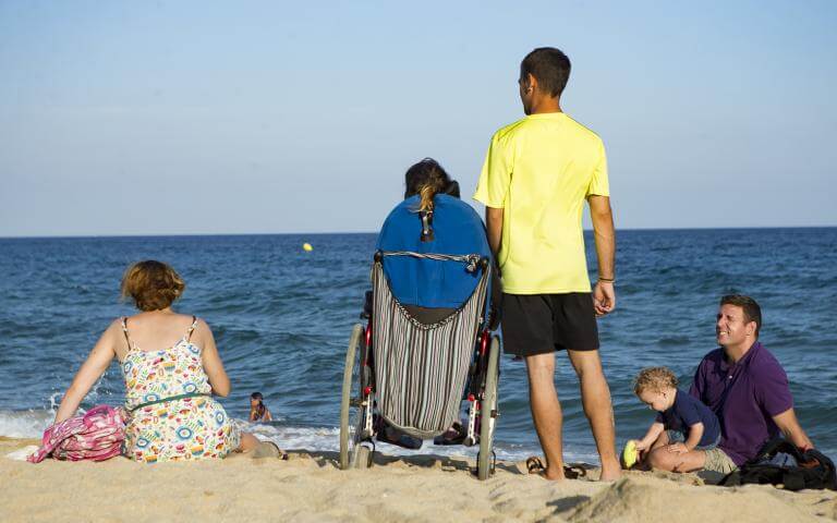 Una persona usuària de cadira de rodes a la platja, acompanyada pel seu assistent personal i altres persones