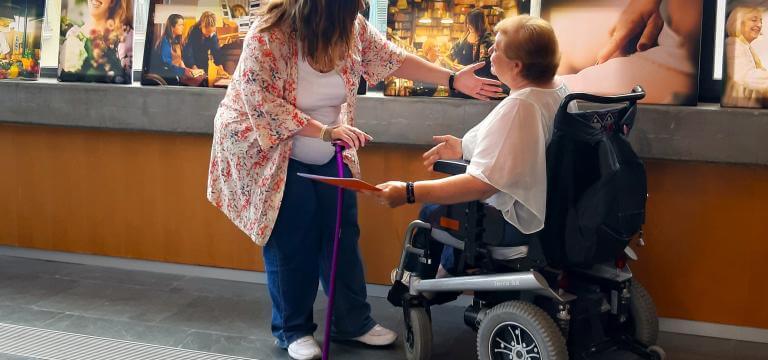 Dos mujeres con discapacidad (una usuaria de silla de ruedas, la otra lleva una muleta) frente a un ventanal y de unas fotografías de la exposición 'Vidas diversas' de ECOM
