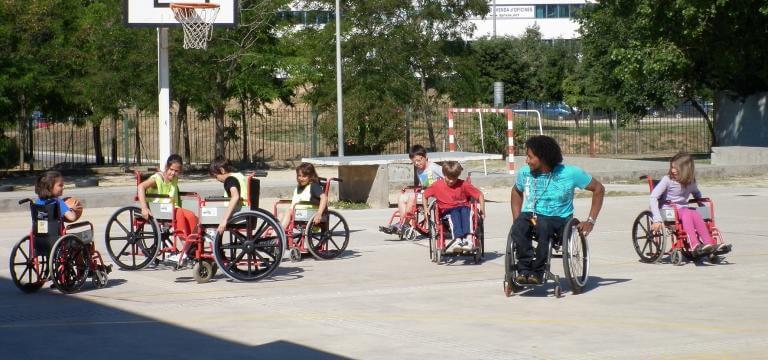 Un grupo de alumnos/as en el patio del colegio, en la cancha de baloncesto, sentados en sillas de ruedas, atendiendo las explicaciones que les está dando un chico con discapacidad (usuario de silla de ruedas) sobre cómo manejar la silla