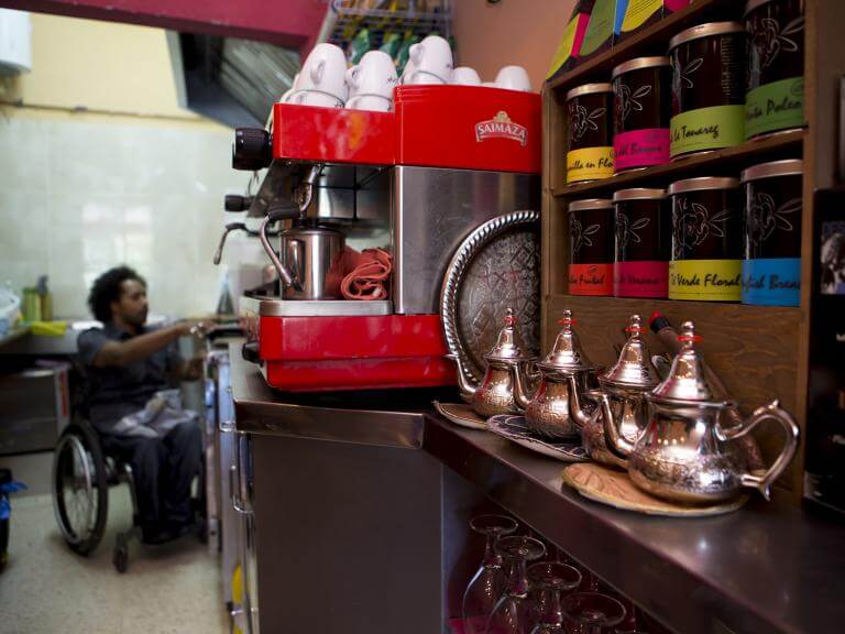 El pla general mostra la banda interior de la barra d'una cafeteria. En primer pla es veuen unes teteres, unes tasses i uns pots, i al fons es veu un noi usuari de cadira de rodes en actitud d'estar servint alguna cosa