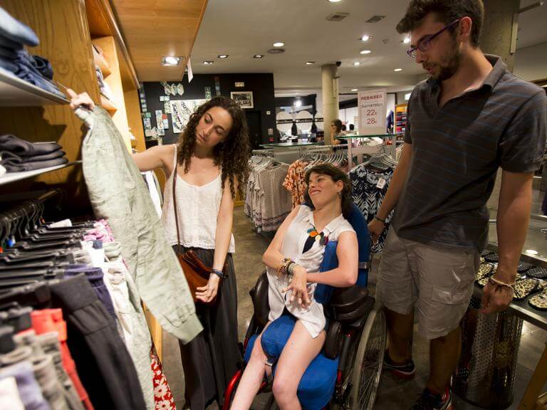 Un noi i dues noies dins una tenda de roba, una d'elles usuària de cadira de rodes. Estant mirant roba, una de les noies li mostra una peça de roba a l'altra (que té una discapacitat) perquè opini i triï. 