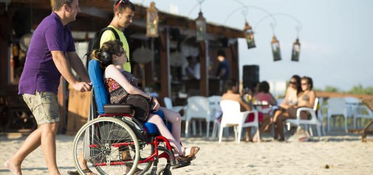 Dos nois acompanyant una noia en cadira de rodes a la platja