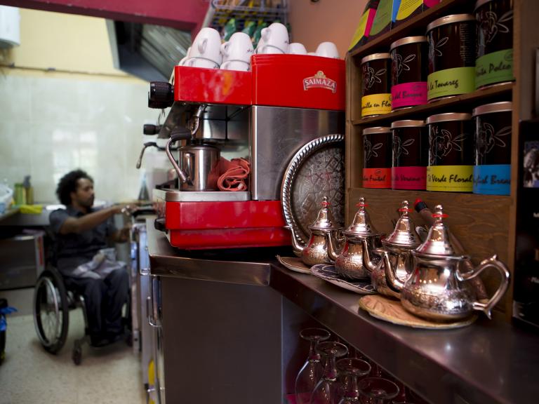 El plano general de la imagen muestra el lado interior de la barra de una cafetería. En primer plano se ven unas teteras, unas tazas y unos botes, y al fondo se ve un chico usuario de silla de ruedas en actitud de estar sirviendo algo