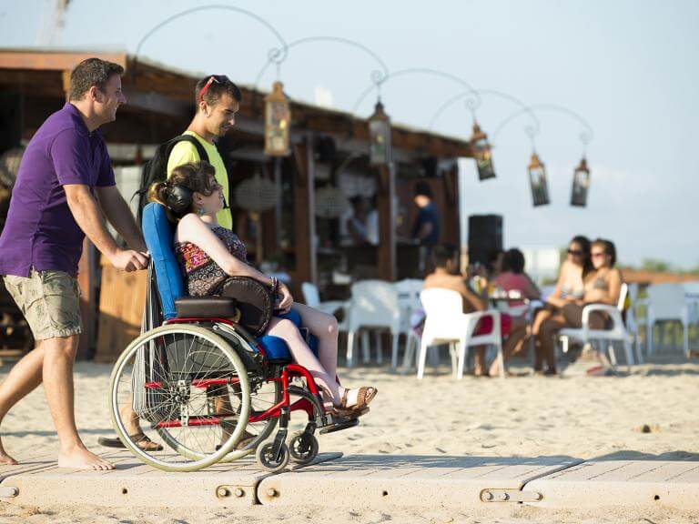 Dos nois i una noia (aquesta amb discapacitat i usuària de cadira de rodes) estan apropant-se al mar avançant sobre la plataforma de fusta que hi ha instal•lada sobre l’arena per facilitar l’accés