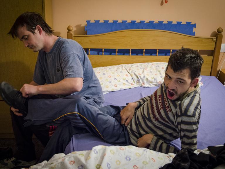 Dos personas sentadas en una cama, una de ellas (que tiene discapacidad) está un poco apoyada atrás mientras la otra le está poniendo los zapatos