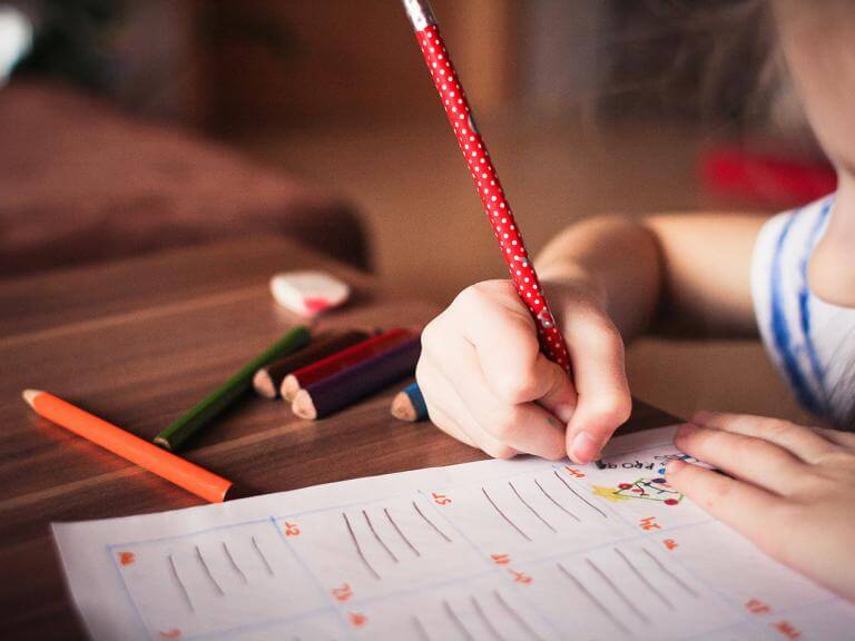 Una niña (de la que sólo se ve un poco el perfil) escribiendo en un papel sobre una mesa donde hay varios lápices