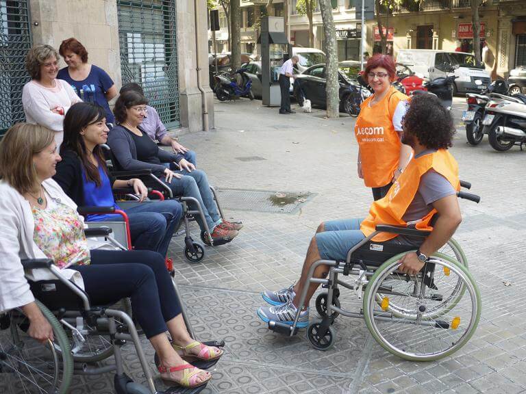 Dos tècnics d'ECOM (un d'ells assegut en una cadira de rodes) dona explicacions a professionals d'una empresa que estan assegudes en cadires de rodes preparades a fer una ruta pel carrer d'una ciutat